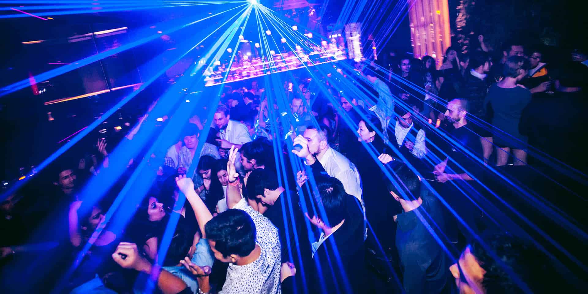 A crowded nightclub with people dancing and socializing under blue laser lights.