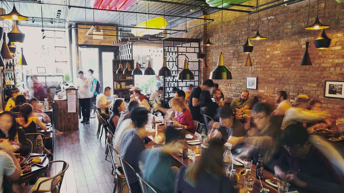Busy restaurant with people dining and interacting at tables, servers attending to guests, and a mix of modern and rustic decor, featuring exposed brick walls and industrial-style lighting.