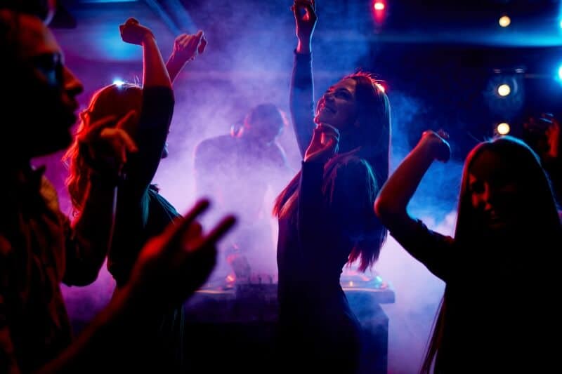 People dancing in a dimly lit club with colorful lights and fog. A DJ is playing music in the background.