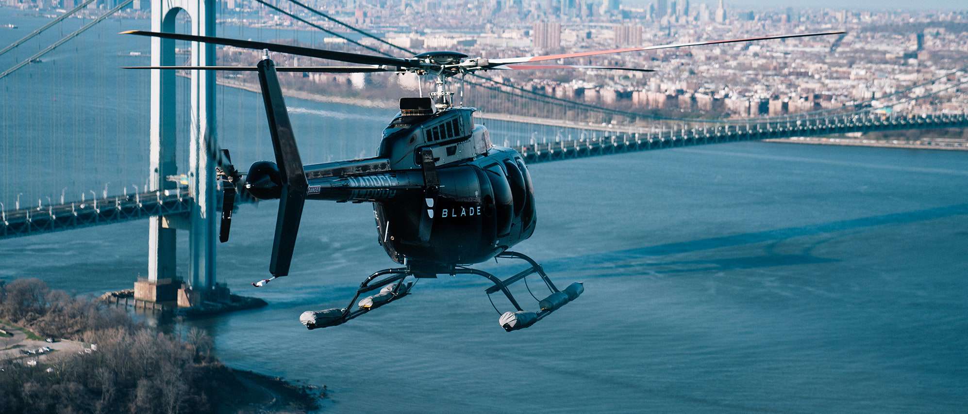 A helicopter with "BLADE" on the side flies over a large body of water with a bridge and cityscape in the background.