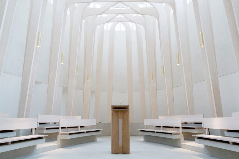 A modern, minimalist chapel interior with light-colored wooden pews and a central wooden lectern, illuminated by natural light from large, vertical windows.