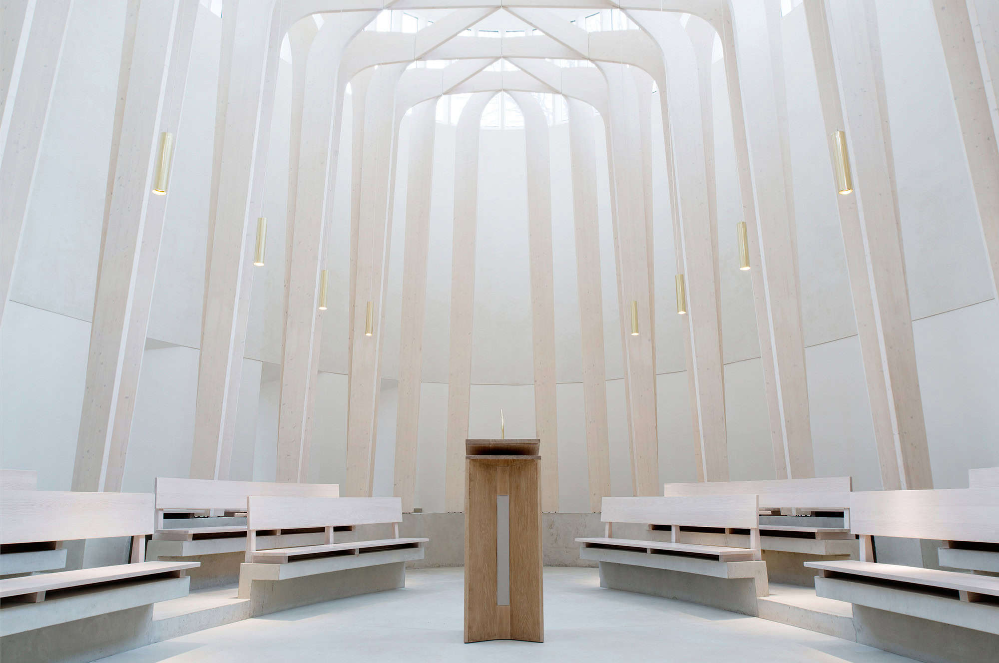 A modern, minimalist chapel interior with light-colored wooden pews and a central wooden lectern, illuminated by natural light from large, vertical windows.