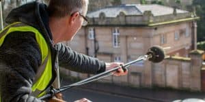 A person wearing a high-visibility vest holds a long microphone boom pole, aiming it towards a building in the background.