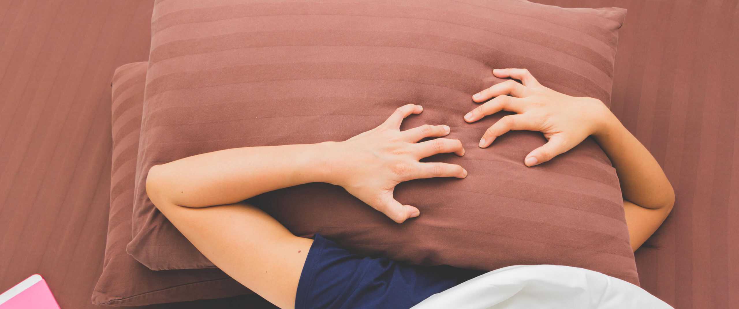 Person lying down with a pillow covering their face and hands gripping the pillow.