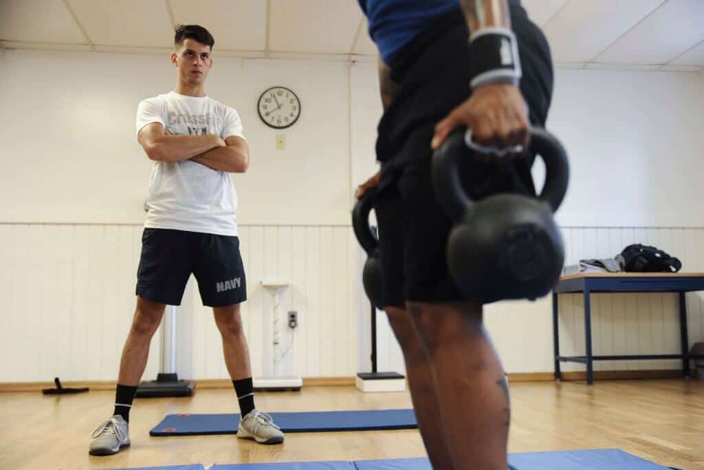 A person in a white "Navy" t-shirt watches another person doing a kettlebell exercise in an indoor gym room.