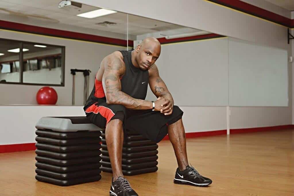 A person in athletic wear sits on stacked exercise steps in an empty fitness studio with a wooden floor and mirrored walls.