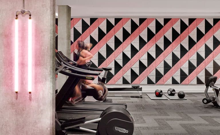 A person runs on a treadmill in a modern gym with a geometric pink, black, and gray wall design. Exercise equipment, including weights and a bike, are visible. Neon lights illuminate the space.