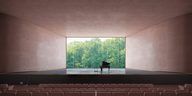 A grand piano on an empty stage in a modern room with pink walls and a large window showing a view of a green forest. Rows of beige seats are in front of the stage.