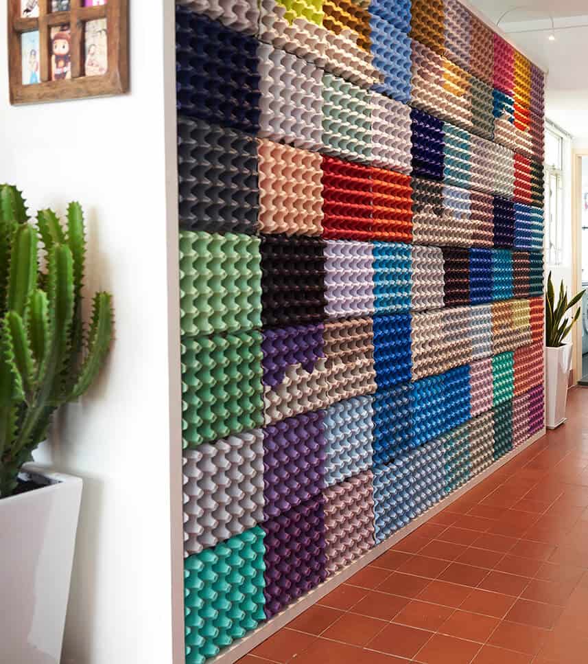 A wall covered in colorful egg cartons arranged in a grid pattern, next to cactus plants in white pots on a tiled floor.