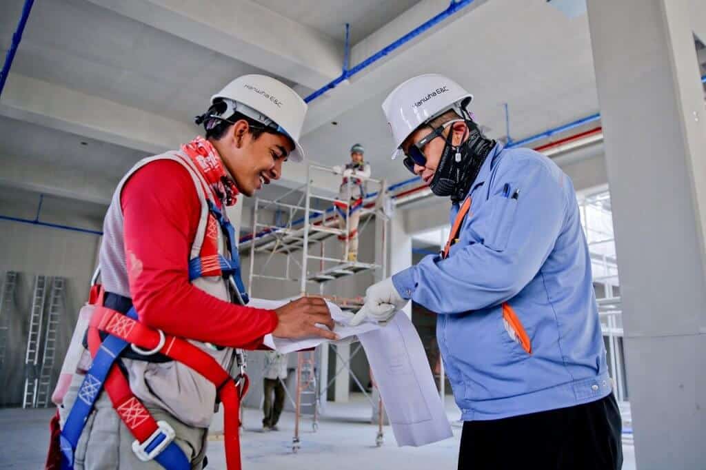 Two construction workers in safety gear and helmets review blueprints at a construction site with scaffolding and ladders in the background.