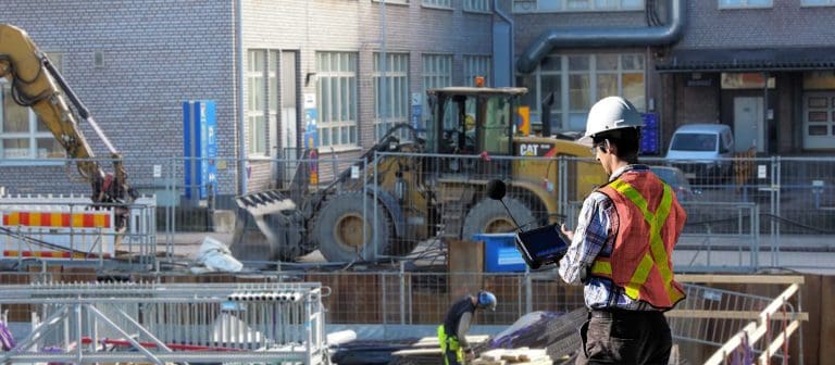 A construction site with heavy machinery. A worker in a reflective vest and hard hat uses a tablet while another worker operates equipment in the background. Buildings are visible behind the site.