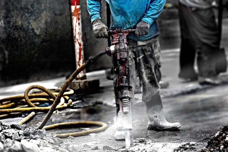 A construction worker wearing gloves and a blue shirt operates a jackhammer on a concrete surface with yellow cables nearby.