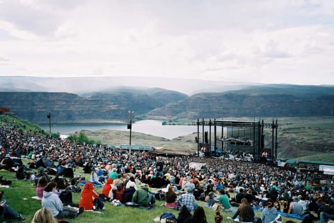 A large outdoor concert surrounded by hills and a lake, with a crowd seated on grassy slopes facing a stage set up with lighting and speakers.