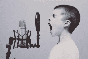 Young child with short hair passionately singing or shouting into a microphone with a pop filter in a monochromatic setting.