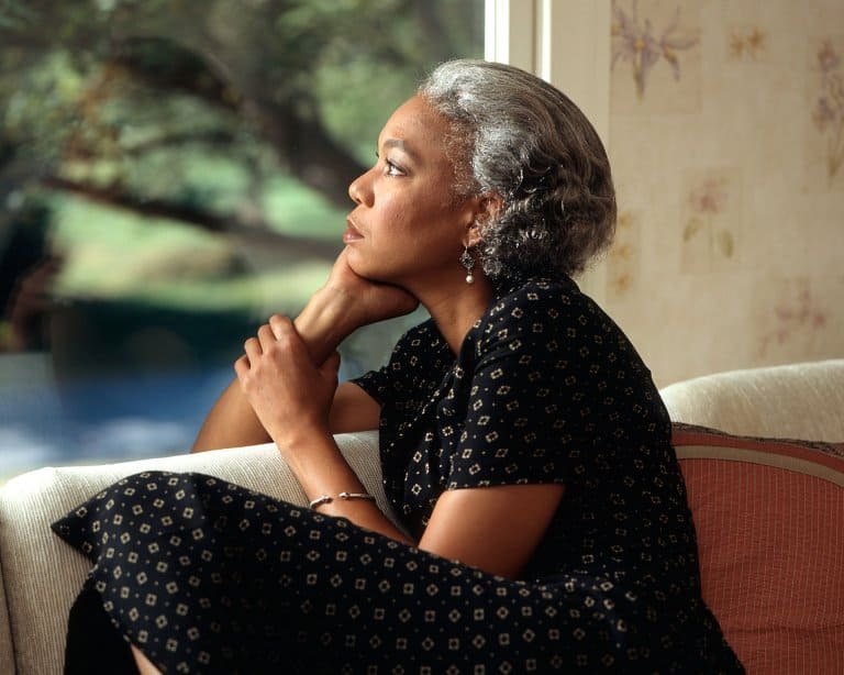 A woman with short gray hair sits on a couch, resting her chin on her hand, and looks thoughtfully out a window. She wears a black patterned dress.