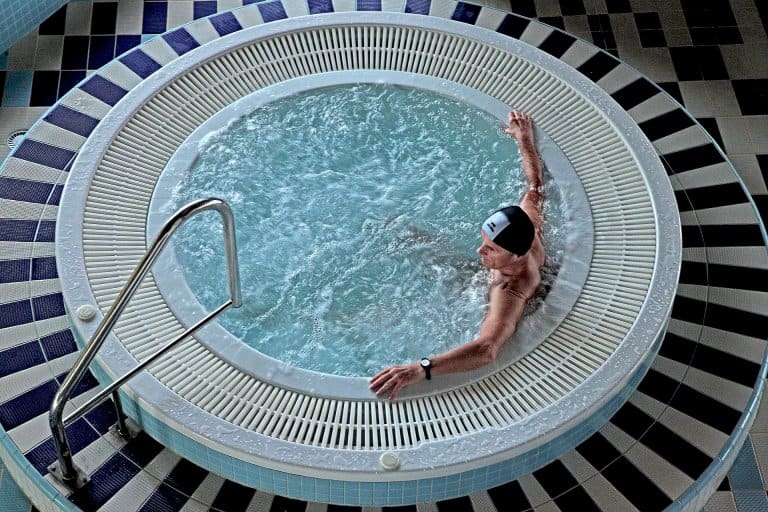 A person wearing a swim cap relaxes in a circular hot tub with swirling water. The hot tub is surrounded by tiles and features a metal handrail on the left.