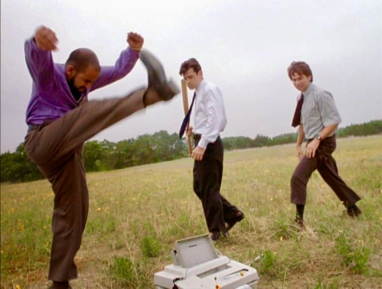 Three men in office attire are outdoors, smashing a printer on the ground; one is kicking it, another is holding a baseball bat, and the third is watching.