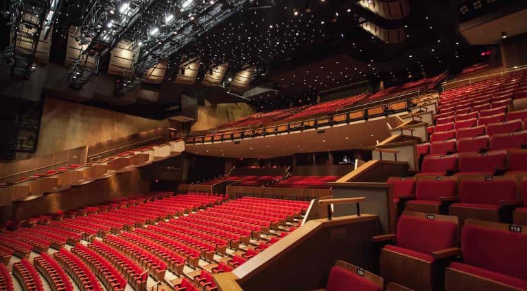 A large, empty theater with red seats and a stage, viewed from the audience area. The ceiling features an array of lights and acoustic panels.