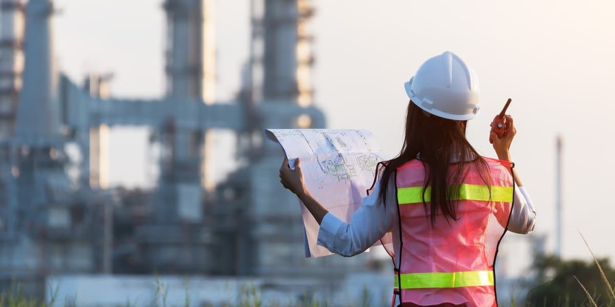A person in a reflective vest and hard hat holds a blueprint and a walkie-talkie, standing in front of an industrial facility.