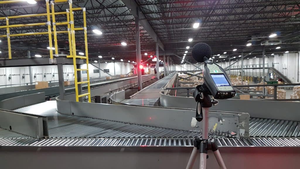 A decibel meter on a tripod is set up inside a large industrial facility with conveyor belts and packages visible in the background.