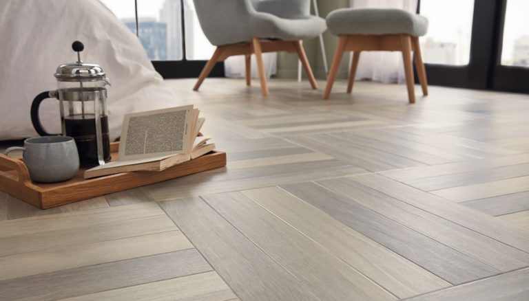 A cozy reading nook with two grey chairs and a wooden tray on the floor holding an open book, a French press coffee maker, and a cup. The floor features a herringbone pattern.