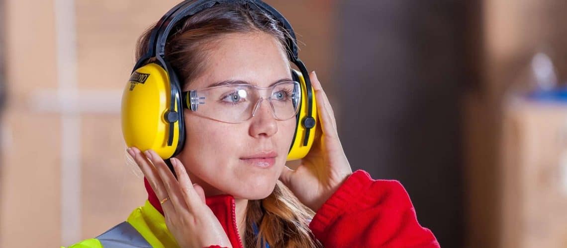 A person wearing safety goggles, ear protection, a high-visibility vest, and a red jacket is adjusting their ear muffs in a workplace setting.