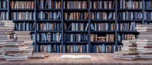 A wooden table with an open book in the middle, surrounded by stacked books, positioned in front of a filled bookshelf.