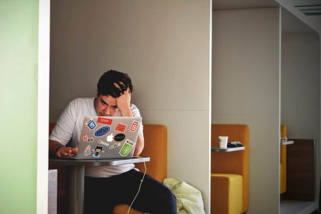 A person with dark hair is sitting at a table in a booth, working on a laptop covered with various stickers, and resting their head on their hand.